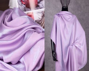 May include: A close-up of a draped, light purple fabric with a shiny, satin-like texture. The fabric is bunched and folded, creating shadows and highlights. A mannequin displays a matching draped garment.
