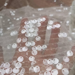 Könnte beinhalten: Nahaufnahme eines durchsichtigen, cremefarbenen Stoffes mit zahlreichen, gleichmäßig verteilten, runden, weißen Pailletten. Der Stoff wird gehalten, wodurch weiche Falten und Schatten entstehen. Die Pailletten fangen das Licht ein und erzeugen einen schimmernden Effekt.