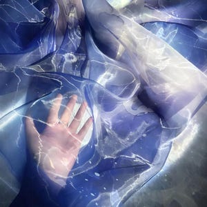 May include: A sheer fabric with a watercolor design in shades of blue and white. The fabric has a flowing, ethereal quality, with light and dark areas creating depth. The design resembles marble or flowing water, with a delicate, translucent appearance.