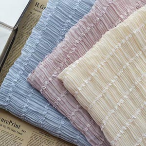 May include: Close-up of four pleated fabrics in pastel colors: light blue, mauve, and cream. Each fabric has a textured, ribbed appearance with a delicate, white floral trim. The fabrics are arranged diagonally, creating a layered effect.