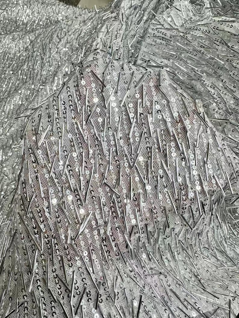 K&ouml;nnte beinhalten: Silberner Stoff mit einer strukturierten, glitzernden Oberfl&auml;che. Der Stoff ist mit kleinen, silbernen Pailletten bedeckt und hat einen fransigen Look.