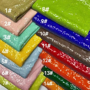 Puede incluir: Una colección de muestras de tela con lentejuelas en varios colores, incluyendo verde oliva, rosa, menta, amarillo y azul claro. Cada muestra está etiquetada con un número, mostrando la gama de colores y texturas disponibles para proyectos de artesanía o diseño.