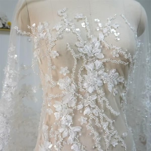 Puede incluir: Una base de tela color nude transparente con intrincados bordados florales blancos, acentuados con pequeñas cuentas blancas, lentejuelas transparentes y pétalos de flores de tela tridimensionales dispuestos en un patrón de ramas y enredaderas.