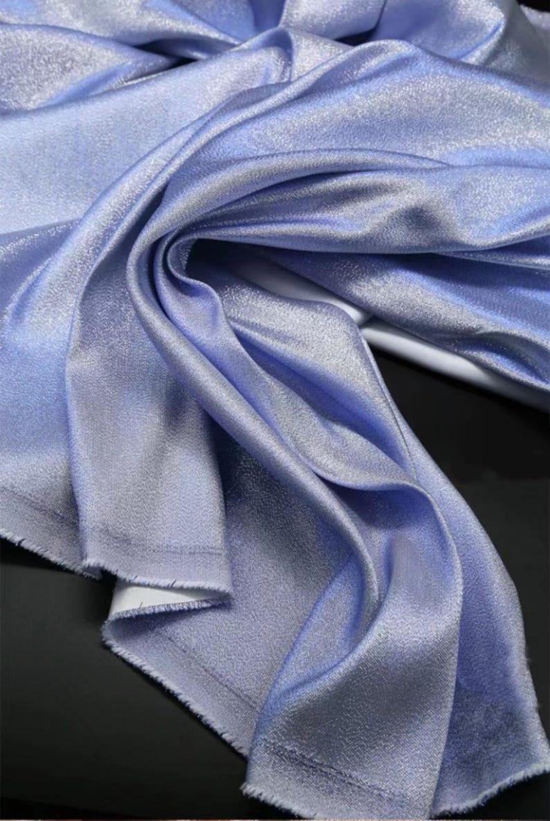 May include: A close-up of a light blue fabric with a subtle shimmer. The fabric is folded and draped, showcasing its soft texture.