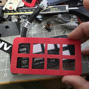 May include: A red switch panel with eight rectangular openings, each designed for a switch. The panel has text labels including "CHASE", "ROCK", "LIGHT BAR", and "FOG". The panel is held in a person's hand, with other electronic components in the background.