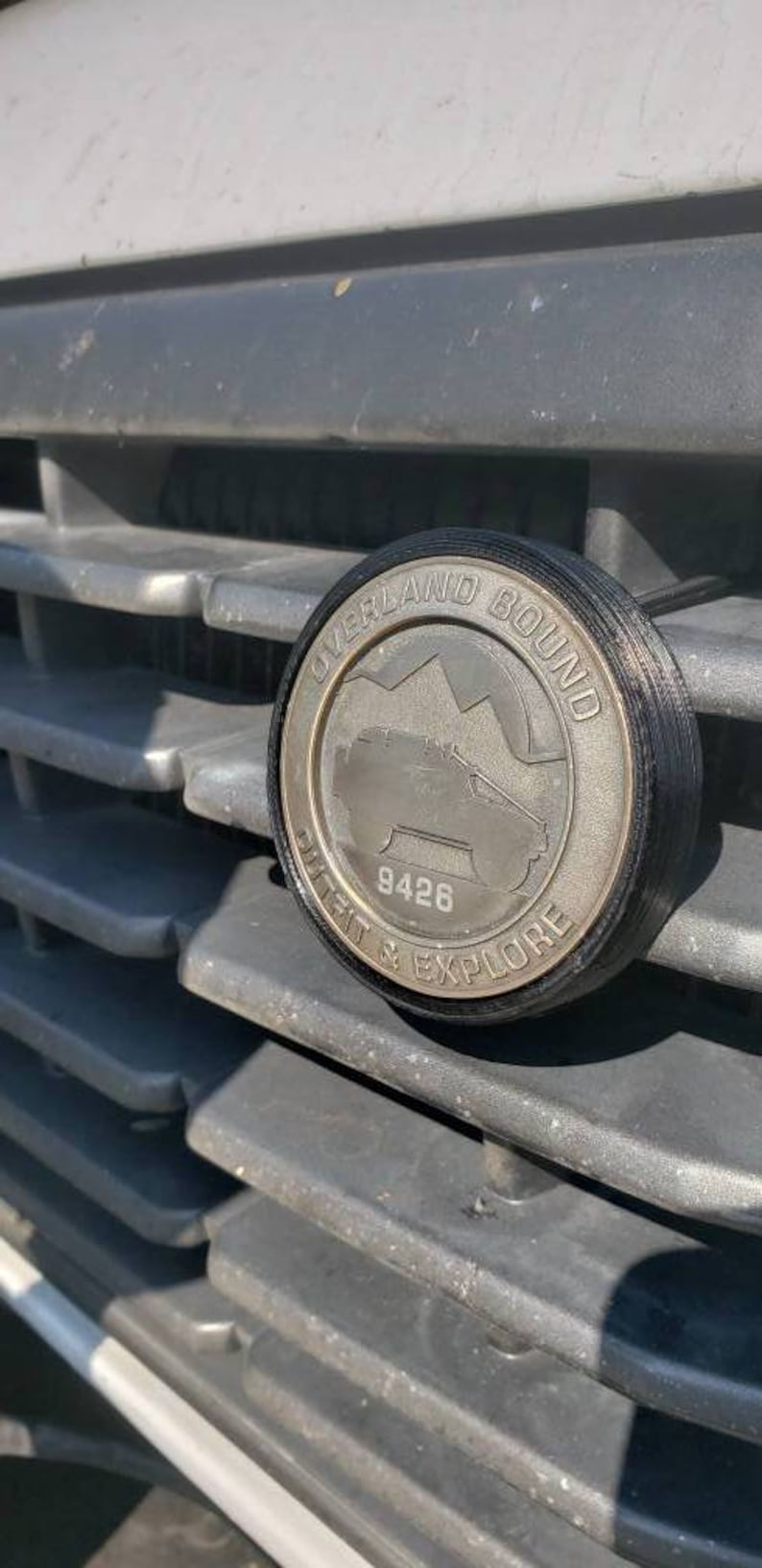 May include: A silver metal emblem with a black border. The emblem features a mountain range and a silhouette of a vehicle with the text "Overland Bound" and "Built & Explore" around the perimeter. The number "9426" is centered on the emblem.