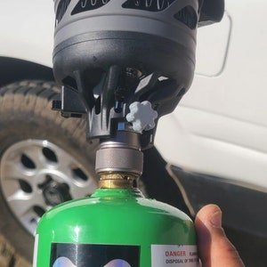 Peut inclure: Une bonbonne de propane verte avec un autocollant noir qui dit "SOLVEFUNCTION" et un réchaud de camping noir et gris fixé sur le dessus. Le réchaud est recouvert d'une housse en tissu noir avec le mot "JETBO" imprimé dessus.