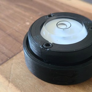 Puede incluir: Un nivel circular negro con un nivel de burbuja transparente en el centro. El nivel est&aacute; hecho de pl&aacute;stico y tiene una superficie texturizada.