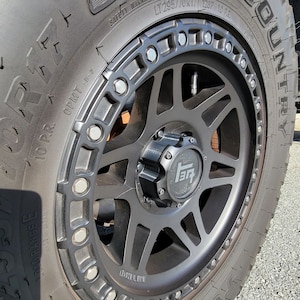 May include: A black tire with a black rim and a black center cap. The tire has the words "Country" and "Off-Road" printed on the side. The rim has a series of bolts around the edge. The center cap has the letters "TR" printed on it.