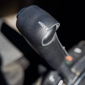 May include: Close-up of a black leather-wrapped control stick, possibly from an aircraft or vehicle. The stick has white stitching and a curved design. The background is blurred, suggesting a focus on the control stick.