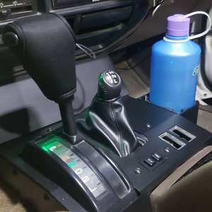 May include: Interior view of a vehicle's console featuring a black gear shift, a smaller black shift lever, and a blue water bottle with a purple cap. The console has a built-in cup holder and USB ports.