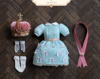 May include: A light blue doll dress with a white collar, pleated bodice, and pink ribbon accents, accompanied by a pink velvet crown, white sheer gloves, white ruffled socks, and a pink ribbon sash labelled BROTHER SHIRT BY SIMON YUEN.