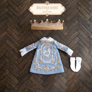 May include: A light blue long-sleeved shirt with white lace trim, gold embroidery, and a central carousel horse motif. The set includes a tan lace crown with pearl accents and a pair of white socks. The text BROTHER SHIRT BY SIMON YUEN is visible.