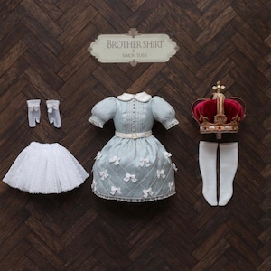 Può includere: Un set di abiti in miniatura su una superficie di legno. Il set comprende un vestito azzurro con fiocchi bianchi, una gonna di pizzo bianco, guanti bianchi, una corona rossa e dorata e calze bianche. Il cartello sopra recita "Brother Shirt" di Simon Yun.