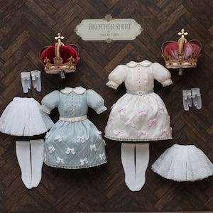 Könnte beinhalten: Eine Auswahl an Puppenkleidung und Accessoires, darunter zwei Kleider, Tütüs, Socken, Handschuhe und Kronen. Ein Kleid ist hellblau mit Schleifen, das andere ist weiß mit rosa Schleifen. Das Schild lautet "Brother Shirt".