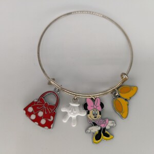 Puede incluir: Un brazalete de plata con cuatro dijes: un bolso rojo con lunares blancos, un guante blanco, una caricatura de Minnie Mouse con un vestido rosa y un par de zapatos amarillos.