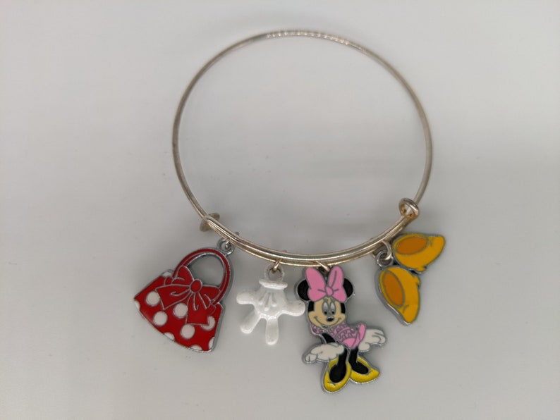 Puede incluir: Un brazalete de plata con cuatro dijes: un bolso rojo con lunares blancos, un guante blanco, una caricatura de Minnie Mouse con un vestido rosa y un par de zapatos amarillos.