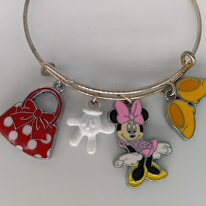 Puede incluir: Un brazalete de plata con cinco dijes: un bolso rojo con lunares blancos y un lazo rojo, un guante blanco, una caricatura de Minnie Mouse con un lazo rosa y dos orejas amarillas de Mickey Mouse.