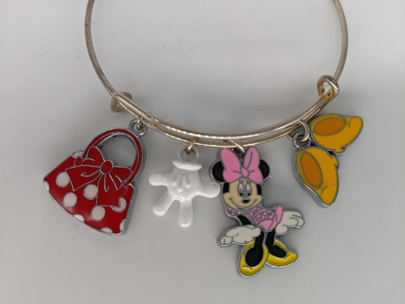 Puede incluir: Un brazalete de plata con cinco dijes: un bolso rojo con lunares blancos y un lazo rojo, un guante blanco, una caricatura de Minnie Mouse con un lazo rosa y dos orejas amarillas de Mickey Mouse.