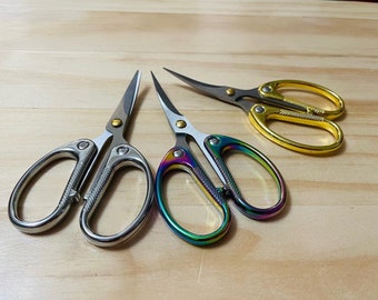 Könnte beinhalten: Drei Paar dekorative Scheren mit gebogenen Klingen. Zwei Paare haben silberne Griffe, eines mit Regenbogen-Finish an den Fingerlöchern. Das dritte Paar hat goldfarbene Griffe. Alle haben silberne Klingen und goldfarbene Nieten.