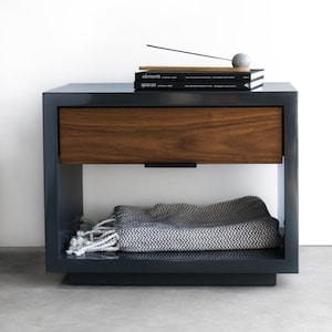 May include: A dark gray nightstand with a wooden drawer and a lower shelf. The nightstand has a stack of books on top, including "elements" and "spaces". A gray ball and a black incense stick are on top of the books.