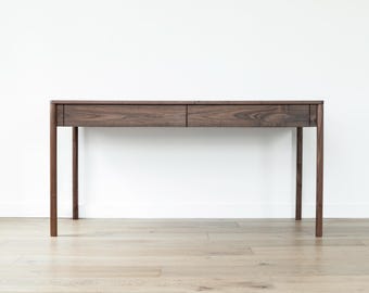 May include: A dark brown wooden desk with two drawers and four slender legs. The desk is made of solid wood and has a simple, modern design. The desk is approximately 60 inches long and 30 inches high.