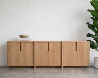 May include: A long, light-toned wooden sideboard featuring six cabinet doors with vertical integrated handles. A small, light-colored bowl and two stacked books titled Kinfolk and Live Beautiful are placed on the left side of the flat top surface.