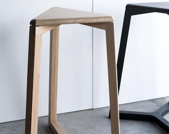 May include: A light-colored wooden stool with a triangular seat and three angled legs. The stool has a minimalist design and is set against a white wall. A black stool is partially visible in the background.