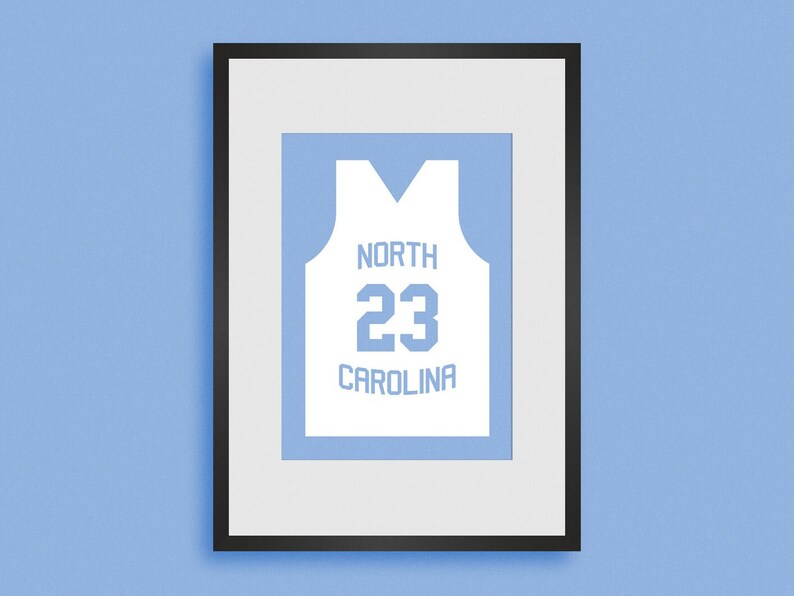 Puede incluir: Una impresi&oacute;n enmarcada de una camiseta de baloncesto blanca con el texto "NORTH CAROLINA" y el n&uacute;mero "23" en azul.