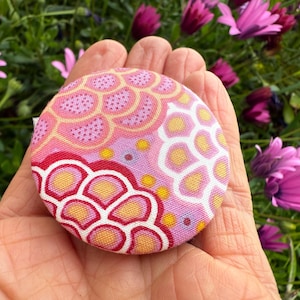 May include: A round, patterned fabric button with a floral design in shades of pink, yellow, and red. The button is held in a hand, with a background of purple flowers and green foliage.