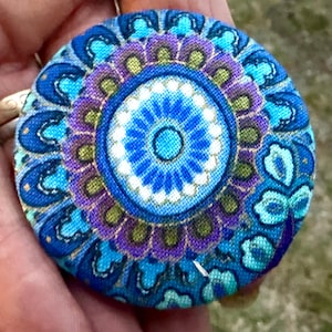 May include: A circular button with a fabric covering, showcasing a mandala-style pattern. The design incorporates shades of blue, teal, purple, and gold in concentric circles. The centre is a bright blue with white detailing.