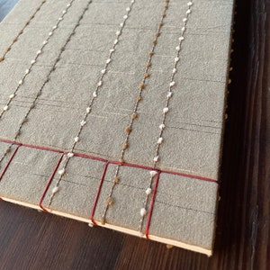 May include: A grey linen journal with red and white stitching. The cover is decorated with rows of brown and white beads.