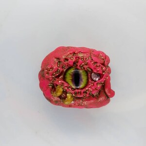 May include: A pink and gold eye with a green iris and a purple pupil. The eye is surrounded by a textured, bumpy surface.