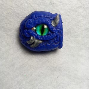 May include: A blue and green dragon eye with a textured surface. The eye is made of polymer clay and has a realistic look.