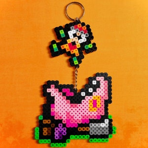 May include: A pink and black pixel art keychain featuring a character from a video game. The character is holding a weapon and is standing on a platform. The keychain is attached to a gold chain.