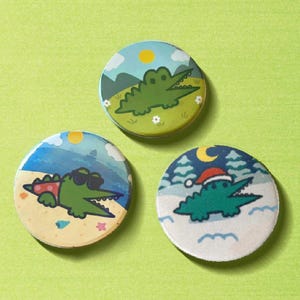 May include: Three round pinback buttons with cartoon alligator designs. One button shows an alligator in sunglasses on a beach, another shows an alligator in a Santa hat, and the third shows an alligator in a landscape.
