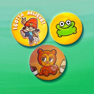May include: Three circular pin-back buttons. One yellow button features a cartoon character in an orange beanie with the text I GOTTA BELIEVE. One yellow button shows a green frog. One orange button depicts a brown bear eating a pink donut.