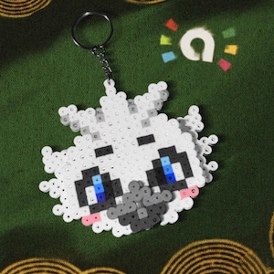 May include: A handmade keychain featuring a pixelated design of a cartoon character's face. The character has white fur, black and gray details, and blue eyes. The keychain is attached to a silver-colored chain and ring.