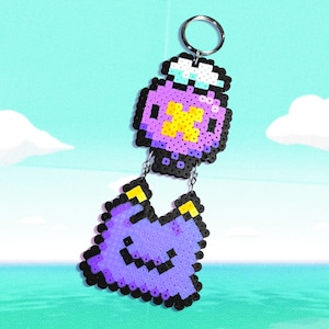 May include: A handmade keychain featuring two pixelated charms. The top charm is a purple potion bottle with a yellow cross and white accents. The bottom charm is a purple ghost with a smiling face and yellow accents. Both charms are connected by a silver chain.