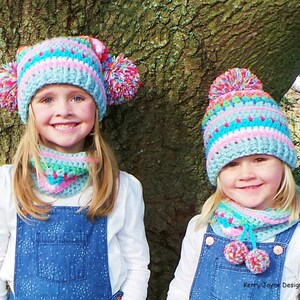 May include: Two girls wearing matching blue and pink striped crochet hats with pom poms and matching blue and pink striped crochet neck warmers. The girls are smiling and standing in front of a tree.