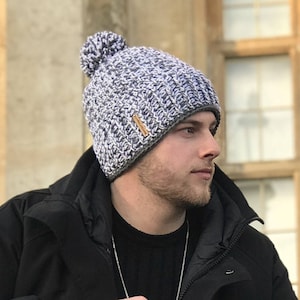 May include: A gray and white knit beanie with a pom pom on top. The beanie has a brown leather tag with the text "Kerry Jayne Designs Patterns" on it.