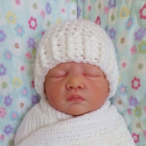 May include: A white crocheted baby hat with a simple stitch pattern. The hat is on a baby wrapped in a white blanket with a floral pattern.