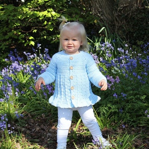 May include: A child in a light blue, crocheted cardigan with wooden buttons and a frilled hem, paired with white leggings and trainers. The background shows greenery and bluebells, typical of a woodland setting.