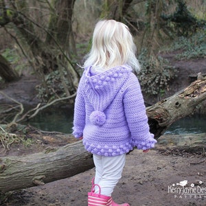 May include: A young girl wearing a purple crocheted hooded sweater with a pom-pom detail. The sweater has a textured design with small bobbles. The girl is standing in a wooded area near a stream.