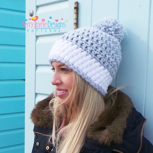 May include: A grey and white crocheted beanie with a white pom pom on top. The beanie has a white band around the bottom. The beanie is being worn by a person with long blonde hair.