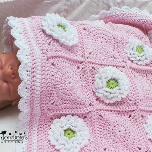 May include: A pink crocheted baby blanket with white and green flowers. The blanket is made with a granny square pattern and has a white crocheted border.