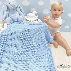 May include: A light blue crochet baby blanket featuring a textured bobble stitch rocking horse design in the center and a matching bobble stitch border. The blanket is displayed alongside a plush blue bunny and a small white rocking horse.