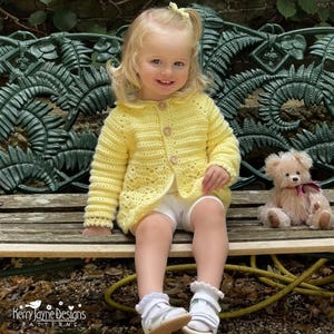 Op de afbeelding: Een kind draagt een gele gehaakte cardigan met houten knopen, een witte korte broek en witte schoenen. Een teddybeer met een roze strik zit naast het kind. De bank staat voor een groene metalen achtergrond. De cardigan is handgemaakt.