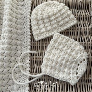 May include: A white, crocheted baby set including a blanket, a beanie-style hat, and a bonnet. The set features a textured, raised stitch pattern. The blanket has a scalloped edge. The bonnet has ties. The set is displayed on a woven surface.