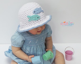 May include: A baby wearing a white crocheted sun hat with blue and green fish appliques. The hat has a white brim and a blue trim. The baby is holding two small plastic buckets.